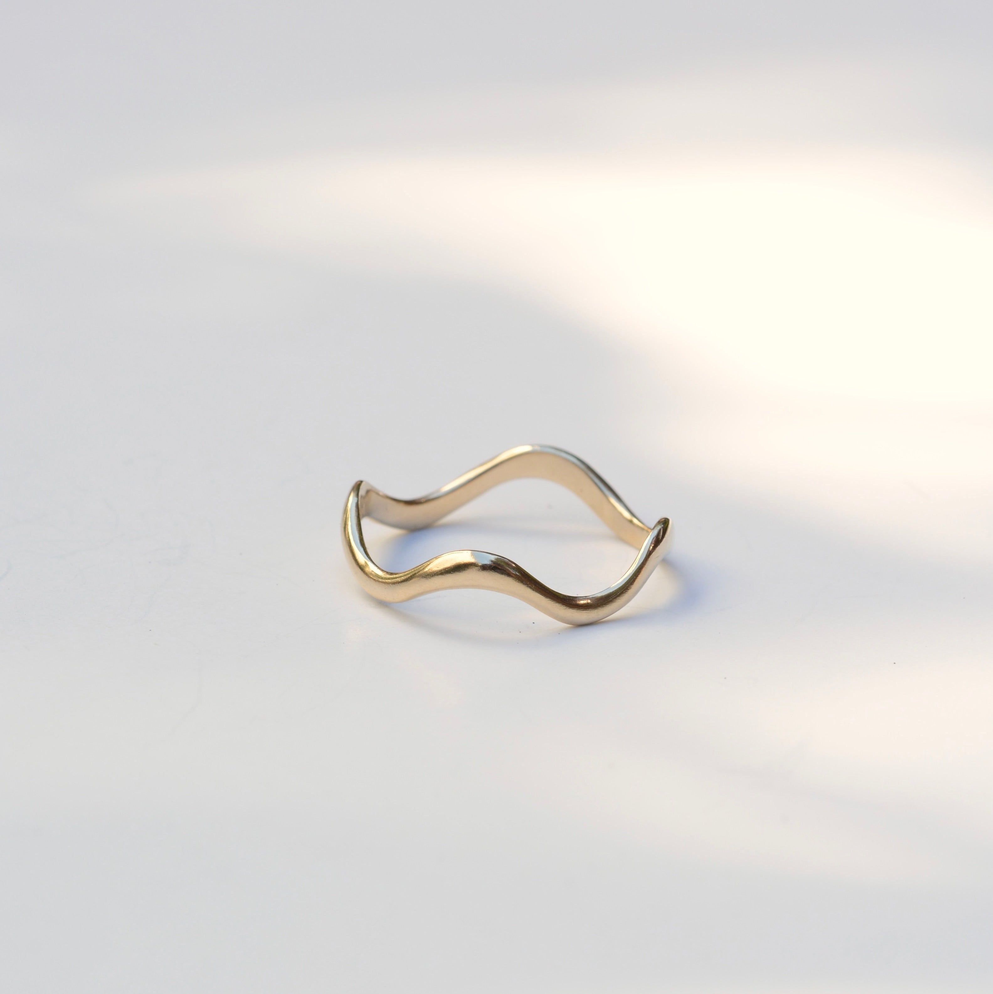A thin, wavy gold ring with an open, flexible design, photographed on a clean white surface with soft, natural shadows.