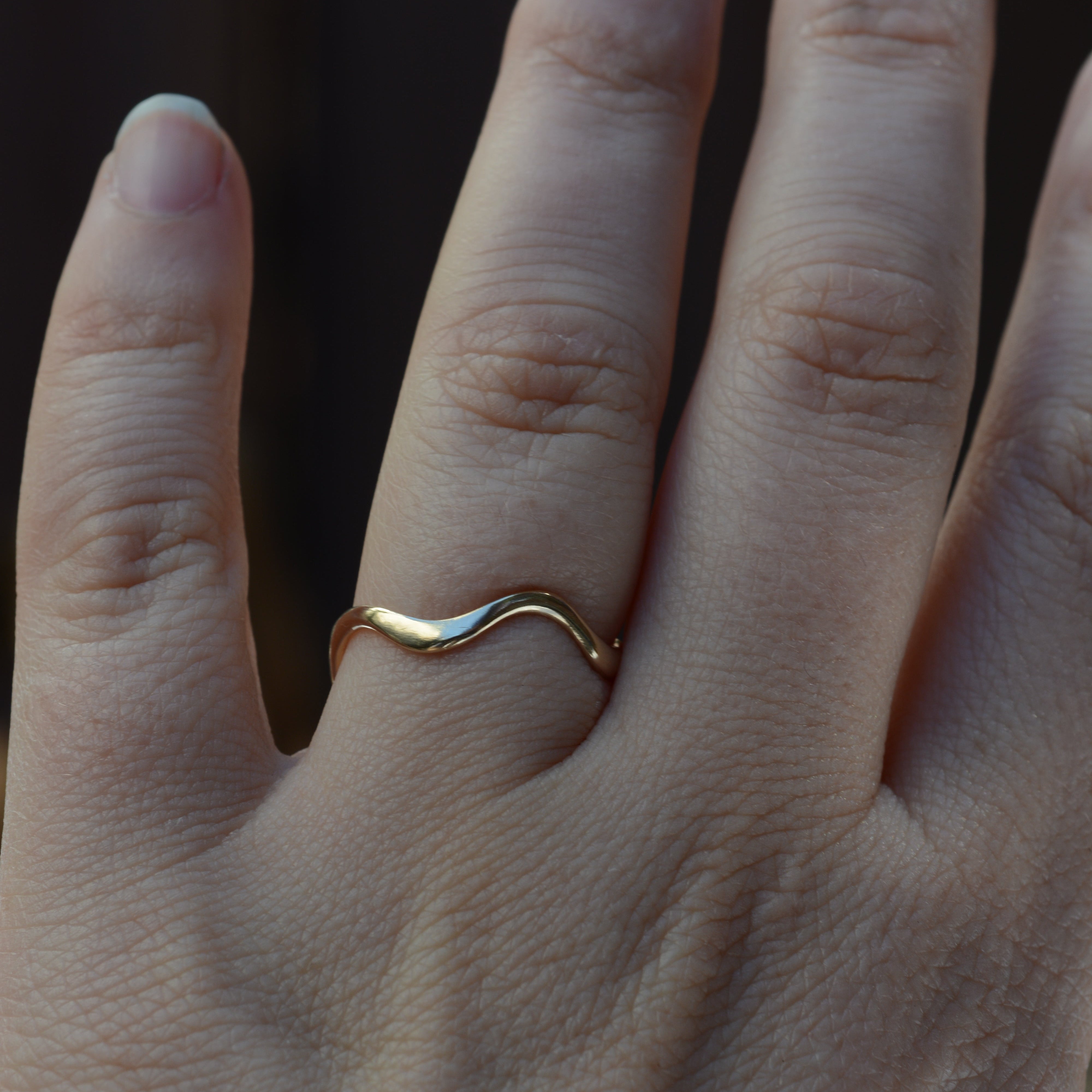 A close-up of a hand wearing a thin, wavy gold band ring on one finger, set against a dark, blurred background.