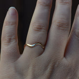 A close-up of a hand wearing a thin, wavy gold band ring on one finger, set against a dark, blurred background.