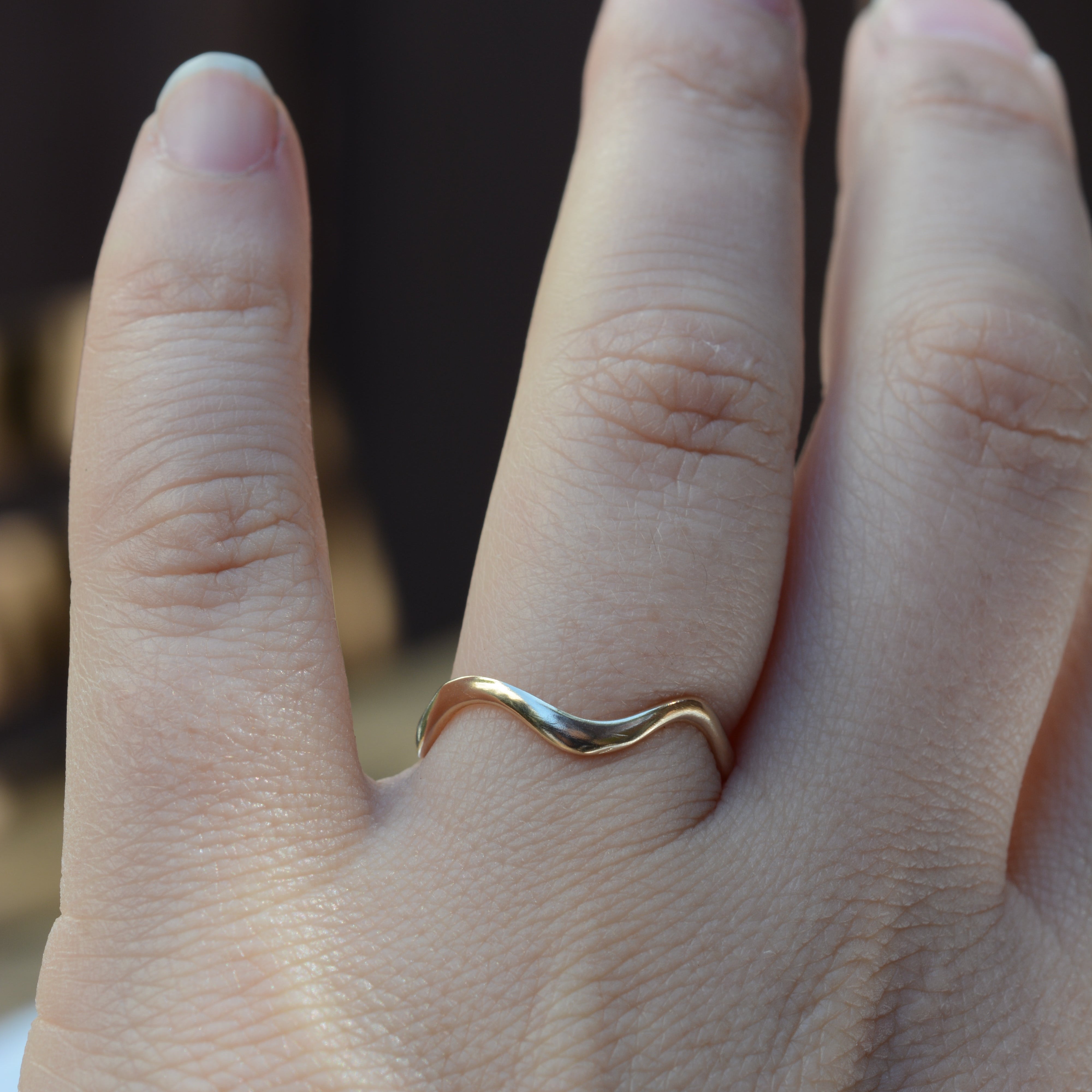 A close-up of a hand wearing a thin, wavy two-tone ring on the ring finger. The ring features one gold-toned half and one silver-toned half, set against a dark, blurred background.