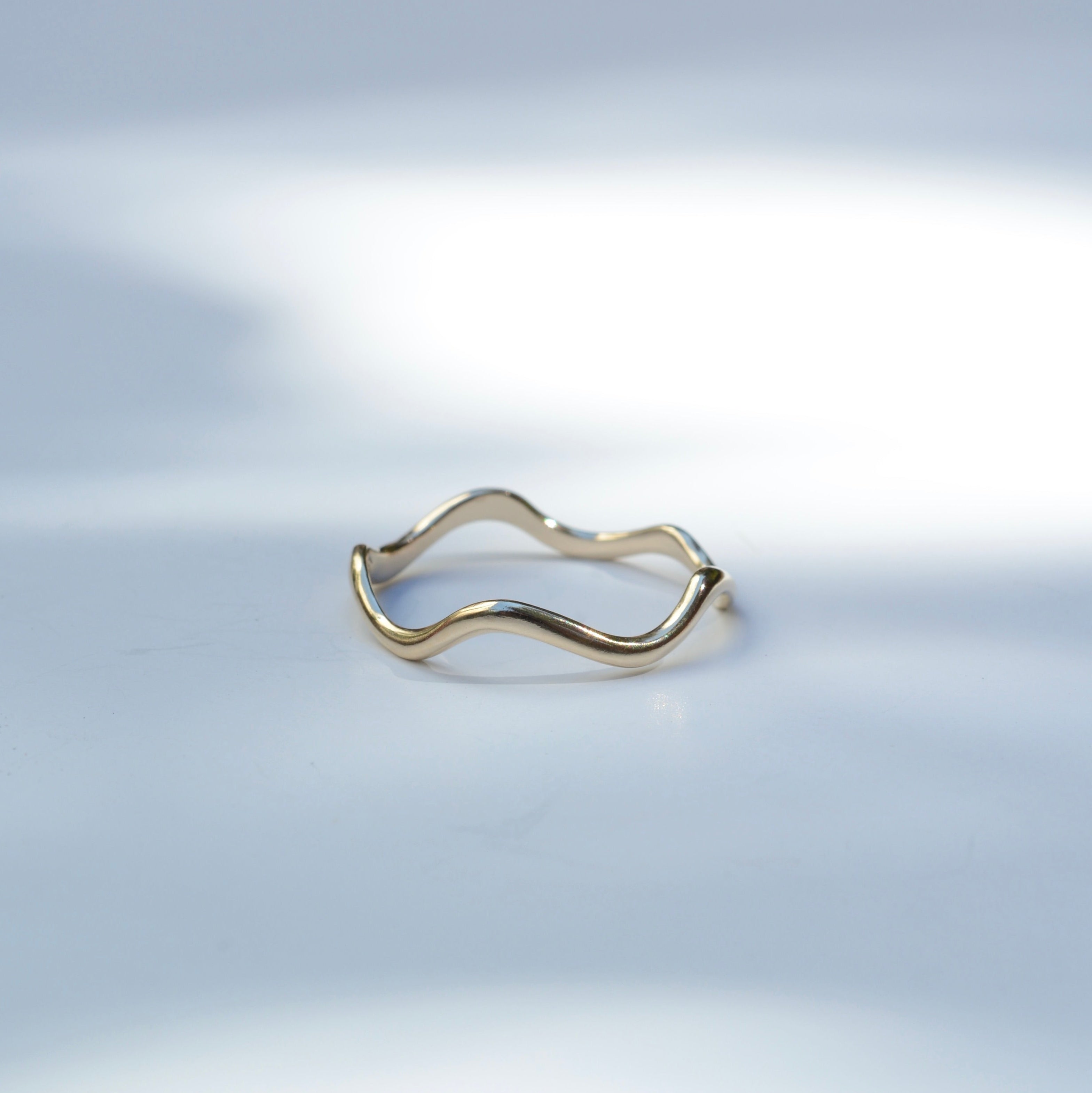 A thin, wavy gold ring with a polished finish, resting on a light gray surface with soft, artistic shadows.
