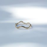 A thin, wavy gold ring with a polished finish, resting on a light gray surface with soft, artistic shadows.