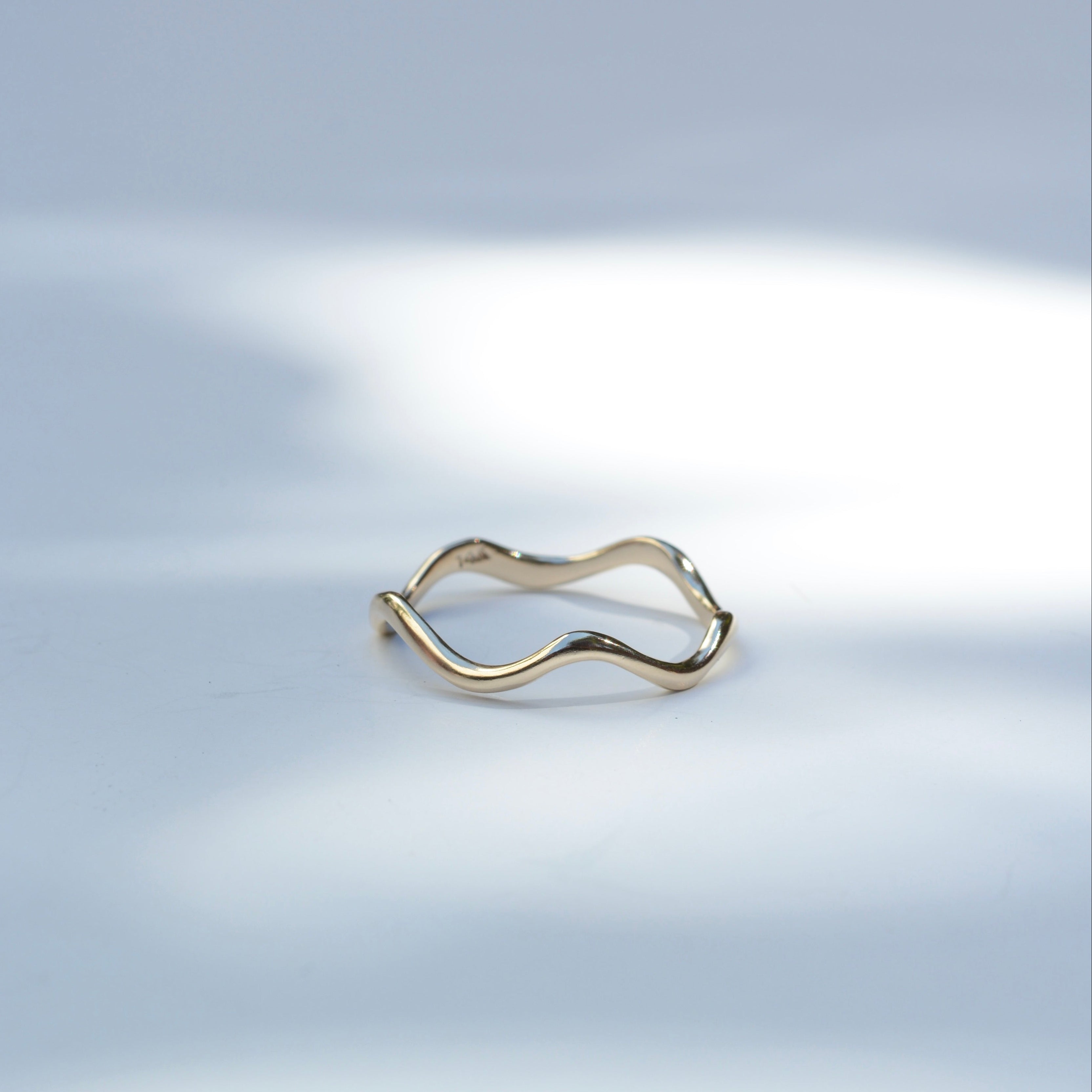 A thin, wavy open-band ring in polished yellow gold, resting on a light gray surface with soft, artistic shadows.