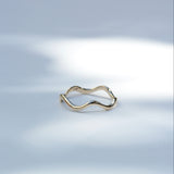 A thin, wavy open-band ring in polished yellow gold, resting on a light gray surface with soft, artistic shadows.