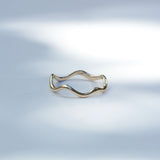 A thin wavy gold ring band photographed on a soft light gray background with a bright blurred light patch behind it.
