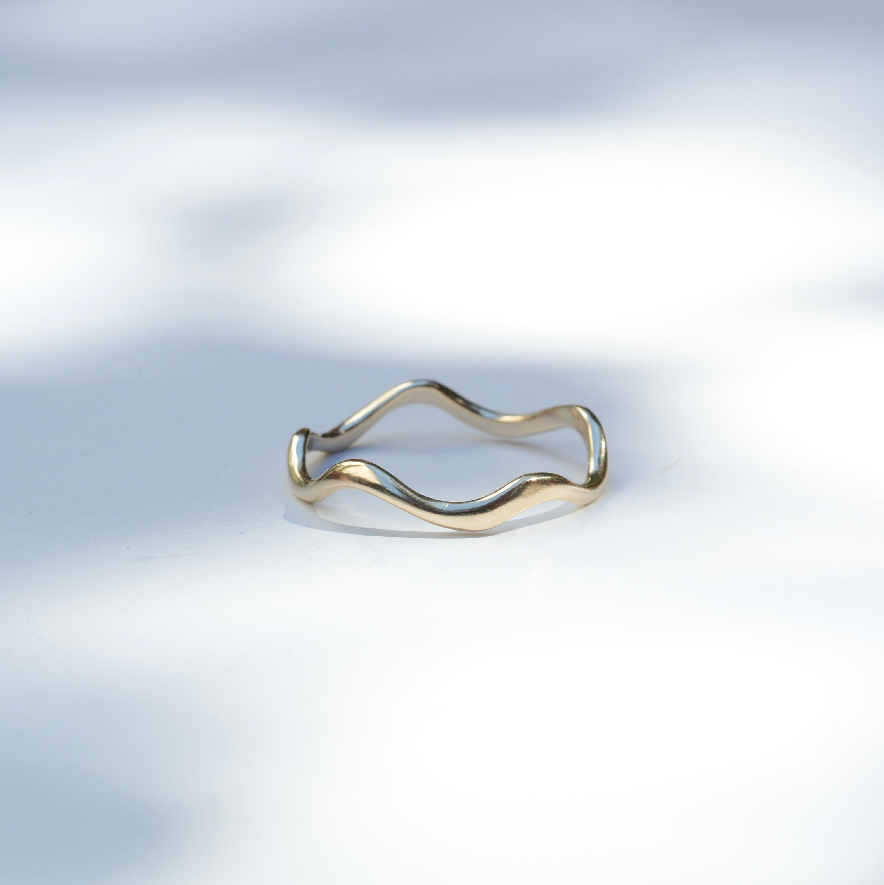 A thin wavy band ring with a two-tone finish, featuring gold on the outer curve and silver on the inner curve, resting on a white surface with soft leaf shadows.