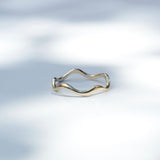 A thin wavy band ring with a two-tone finish, featuring gold on the outer curve and silver on the inner curve, resting on a white surface with soft leaf shadows.