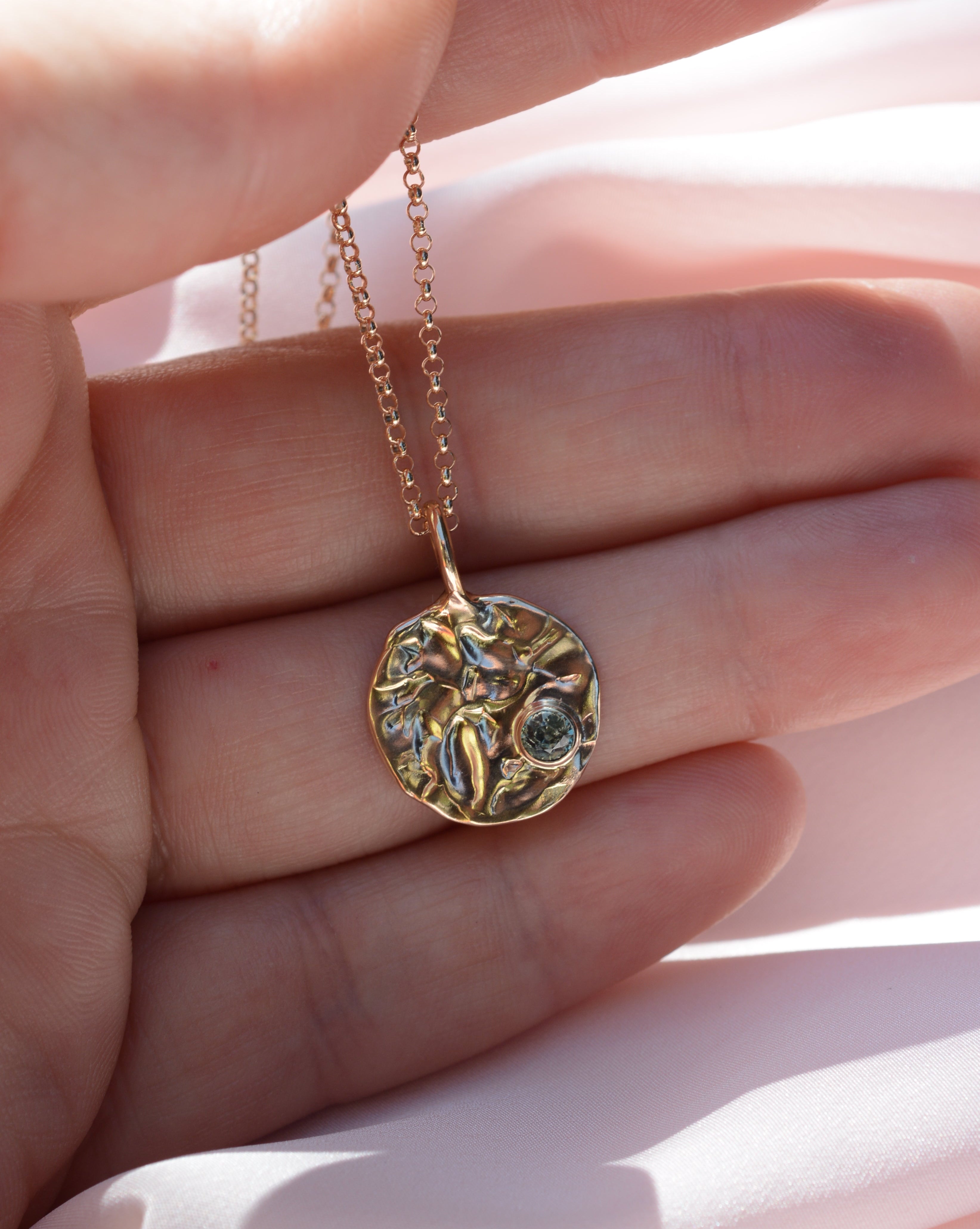 Hand holding a round textured pendant with a light blue sapphire 