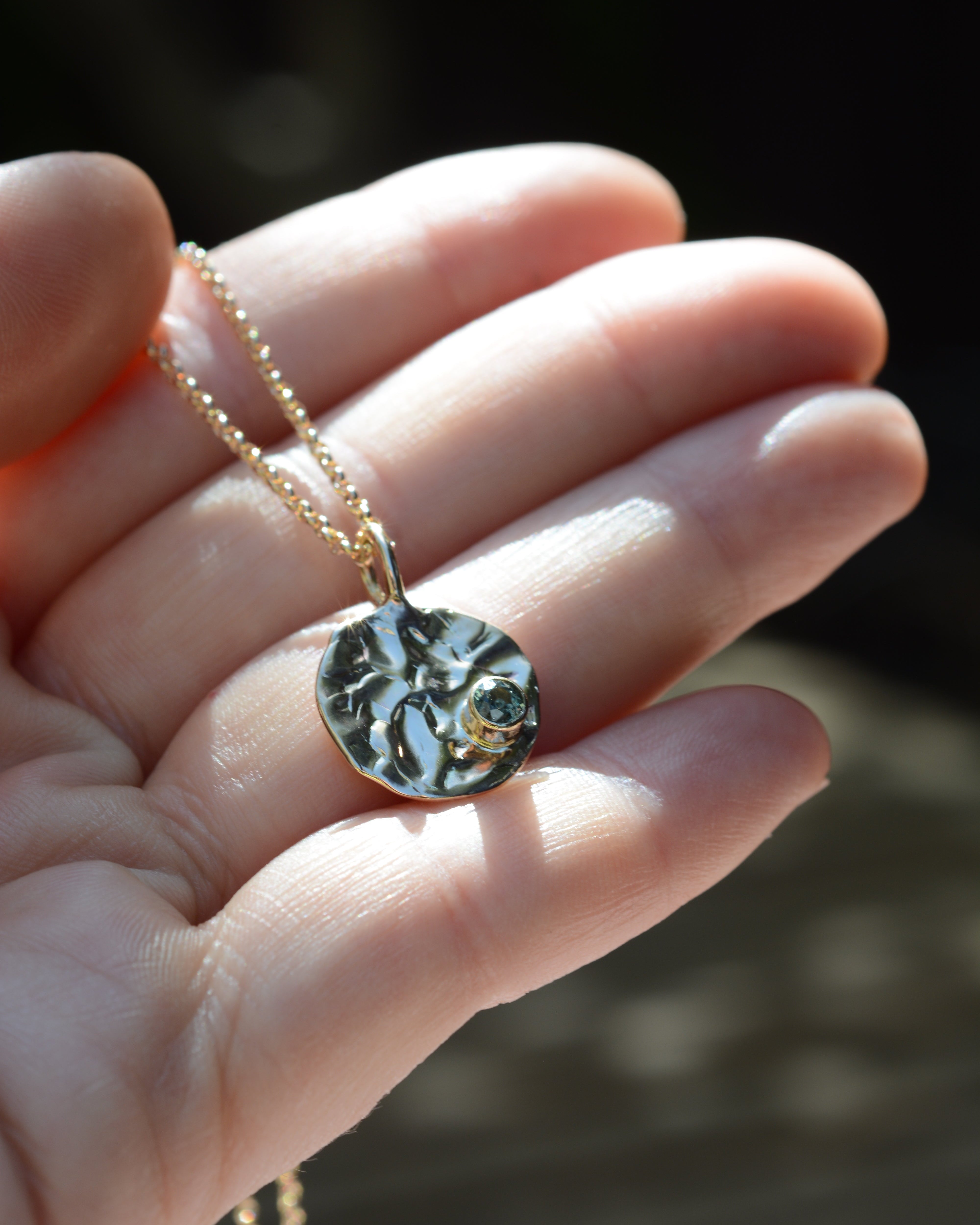 Hand holding a textured round pendant on a chain with a blue sapphire
