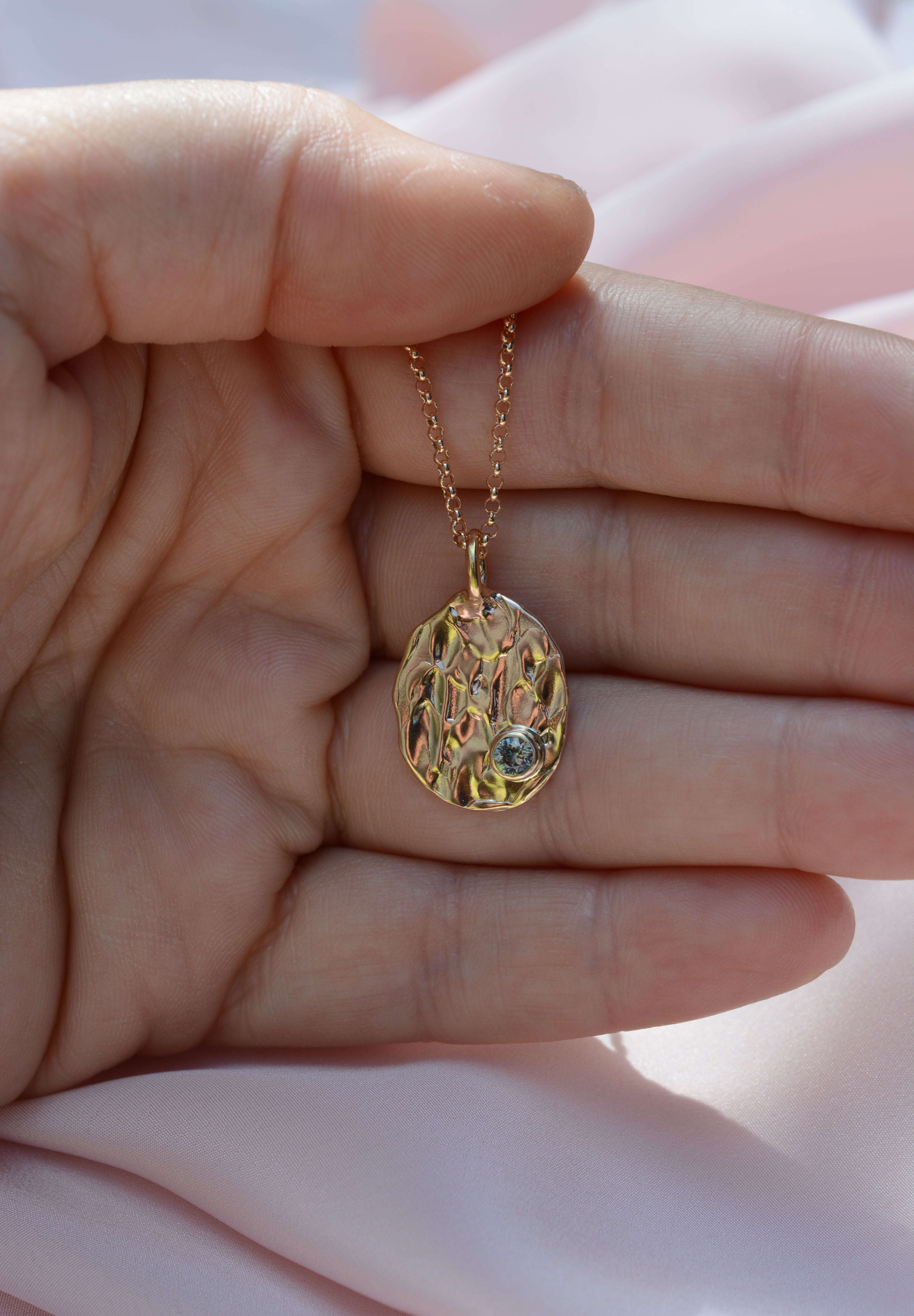 Hand holding an oval pendant on a chain. Pendant has an organic molten texture and a blue sapphire on the bottom right. 