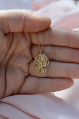Hand holding an oval pendant on a chain. Pendant has an organic molten texture and a blue sapphire on the bottom right. 