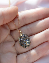 Hand holding an oval pendant with a molten texture and blue sapphire on the bottom right 