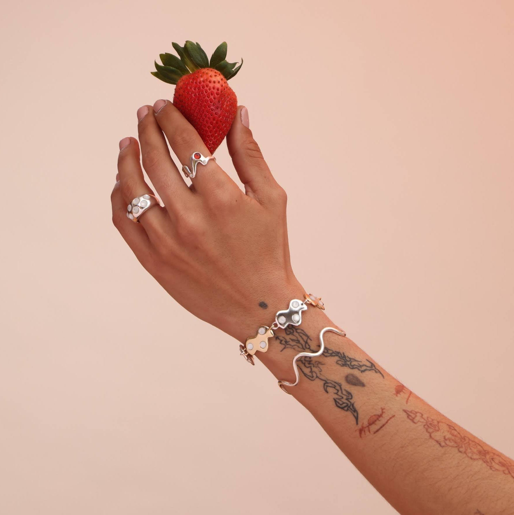Hand holding a strawberry against a beige background with silver jewelry including a silver squiggly cuff