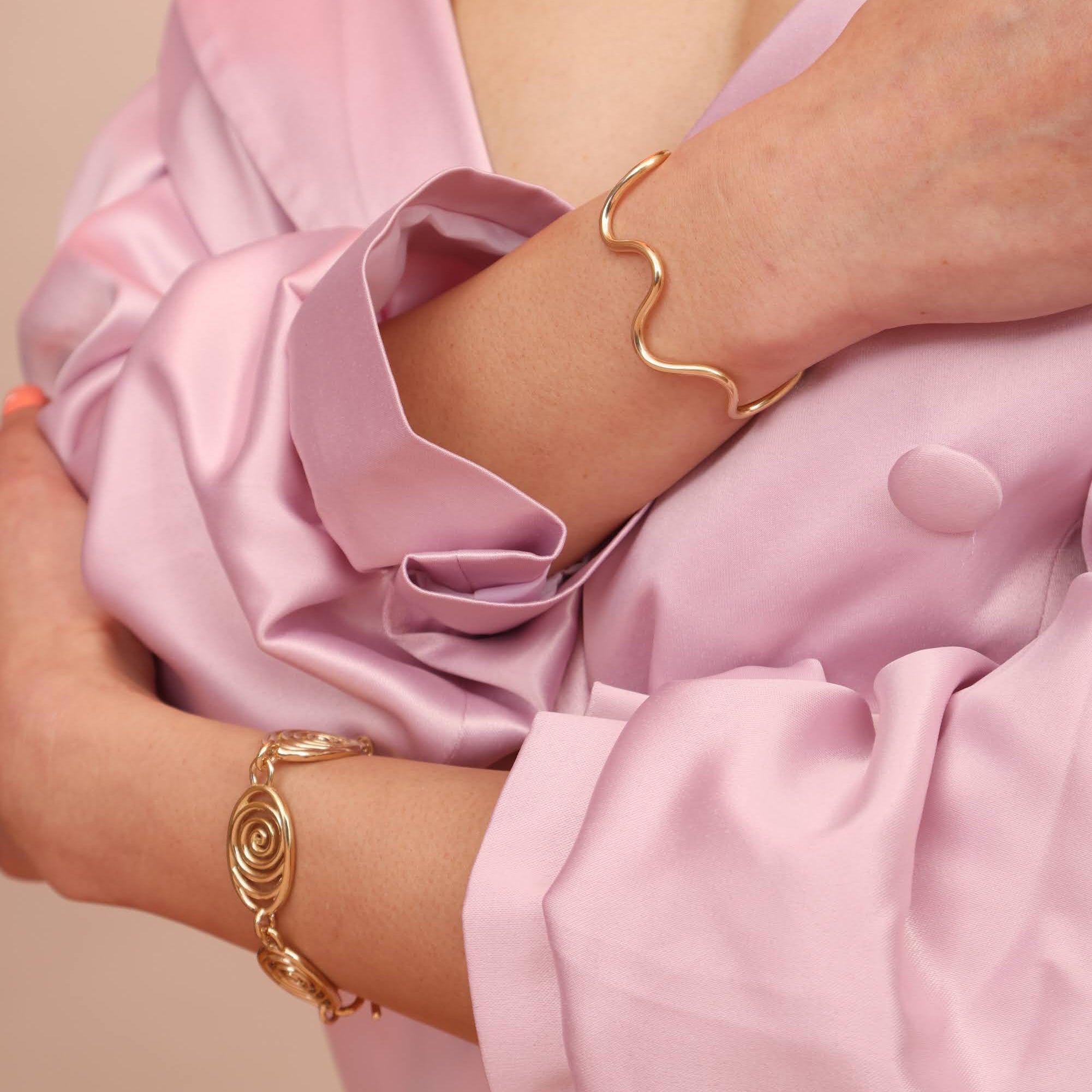 Person wearing gold jewelry including a brass squiggly cuff and a spiral link bracelet on a pink garment with a neutral background