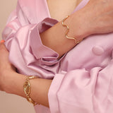 Person wearing gold jewelry including a brass squiggly cuff and a spiral link bracelet on a pink garment with a neutral background