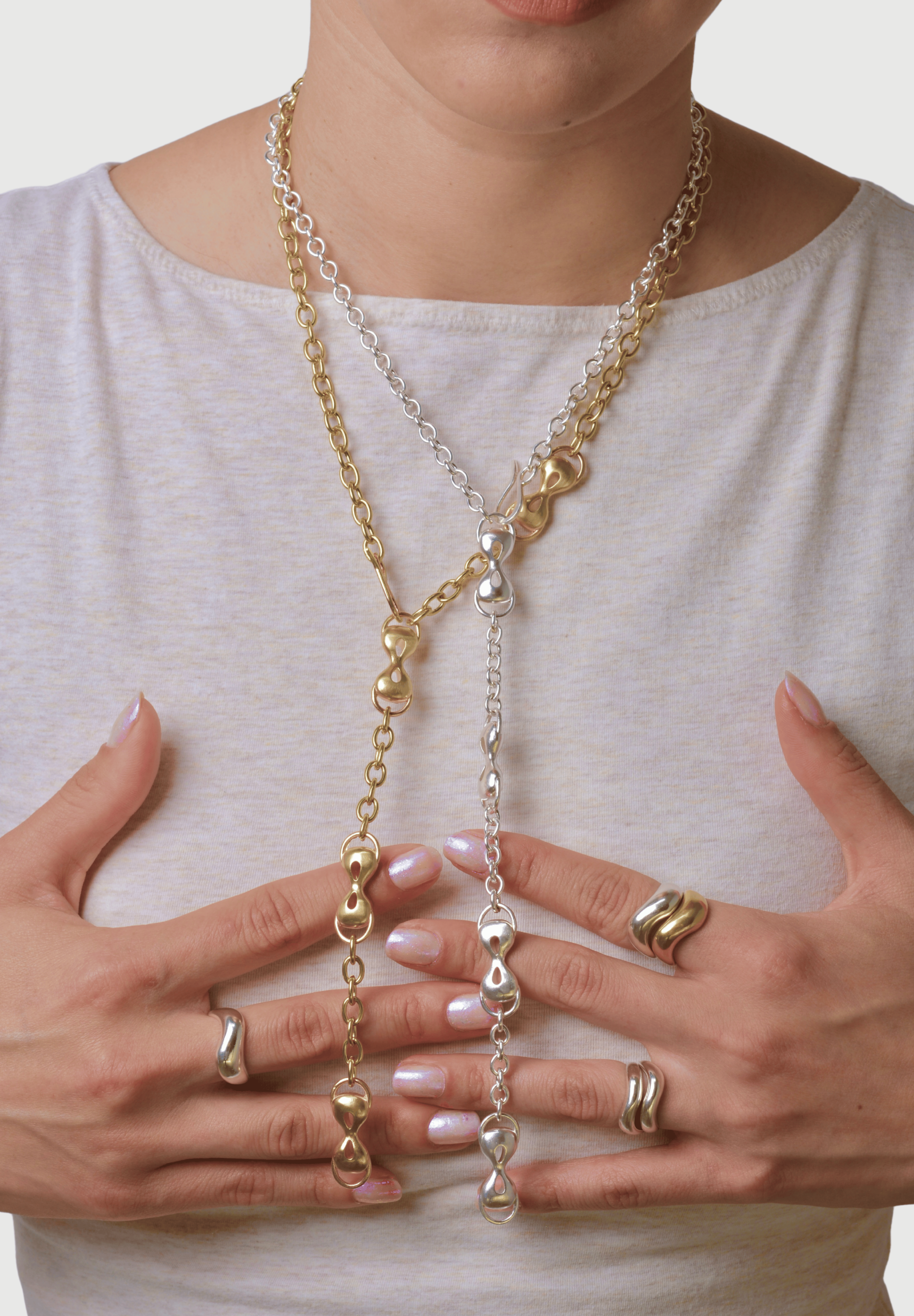 Person wearing two long necklace with bronze and silver chains, holding it with both hands that have wavy stacked rings on them.