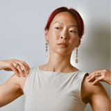 Woman wearing touching her shoulders modeling earrings that are bronze dangles with coral shaped charms and a pearl wearing a white sleeveless top against a plain background